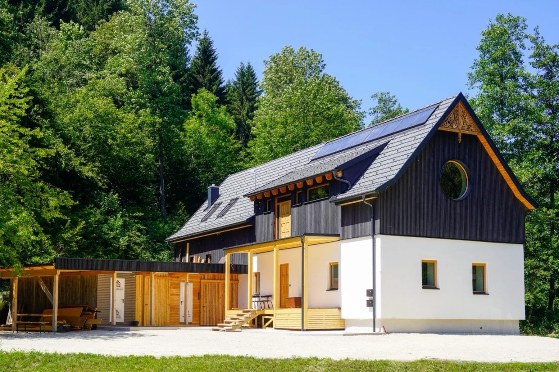 Sanierte Mühle im Tal: weißer Sockel, dunkle Holzschalung im Giebel mit Rundfenster, neue Veranda aus Holz, Wald dahinter.